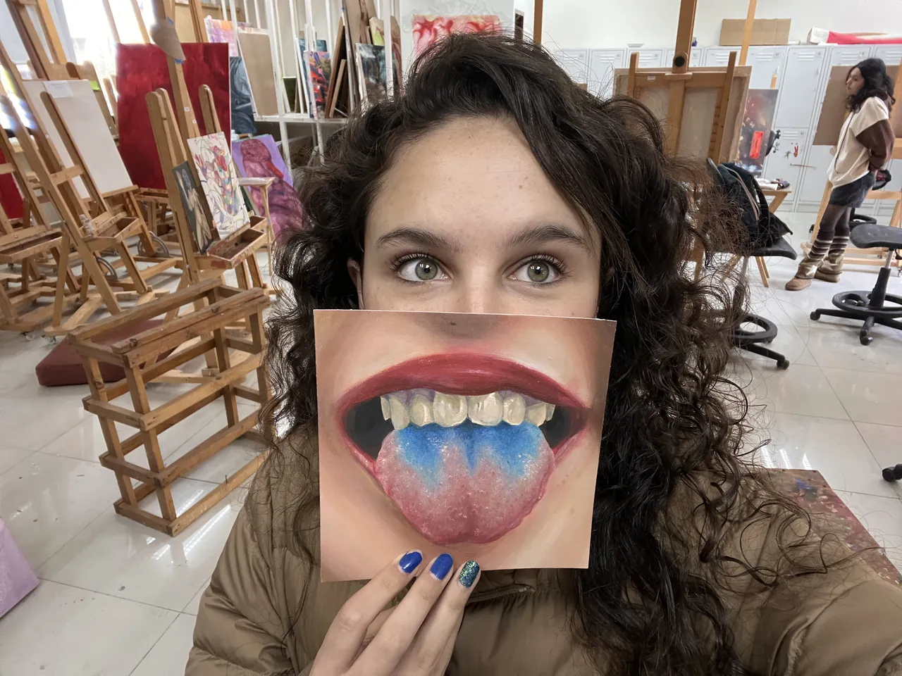 Ximena en el taller sosteniendo una pintura de la serie Bleh frente a su cara.